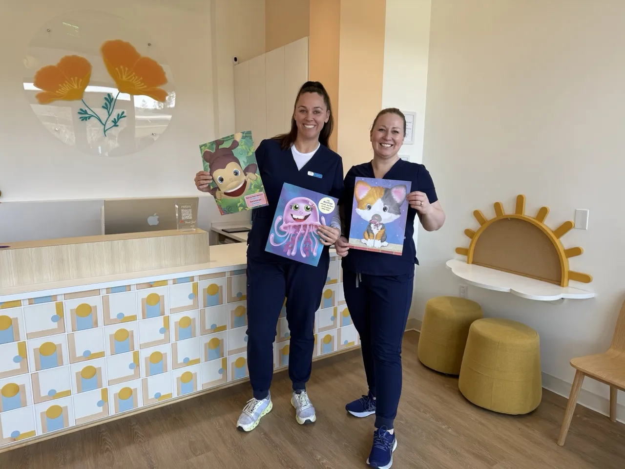Two dental team members, dressed in navy scrubs, happily display colorful children's books in a modern pediatric dental office with a bright and playful design featuring a poppy-themed logo and sun-shaped decor.