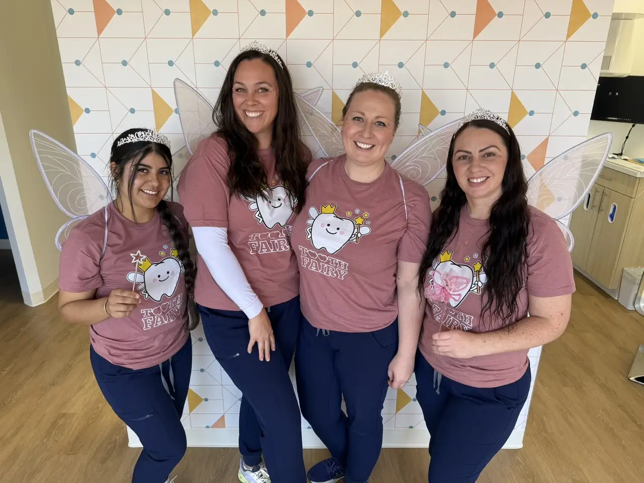 Four team members from Poppy Kids Pediatric Dentistry pose together dressed as tooth fairies, wearing matching pink shirts with a smiling tooth graphic, tiaras, and fairy wings. They hold wands and stand in front of a colorful geometric wall, smiling brightly.