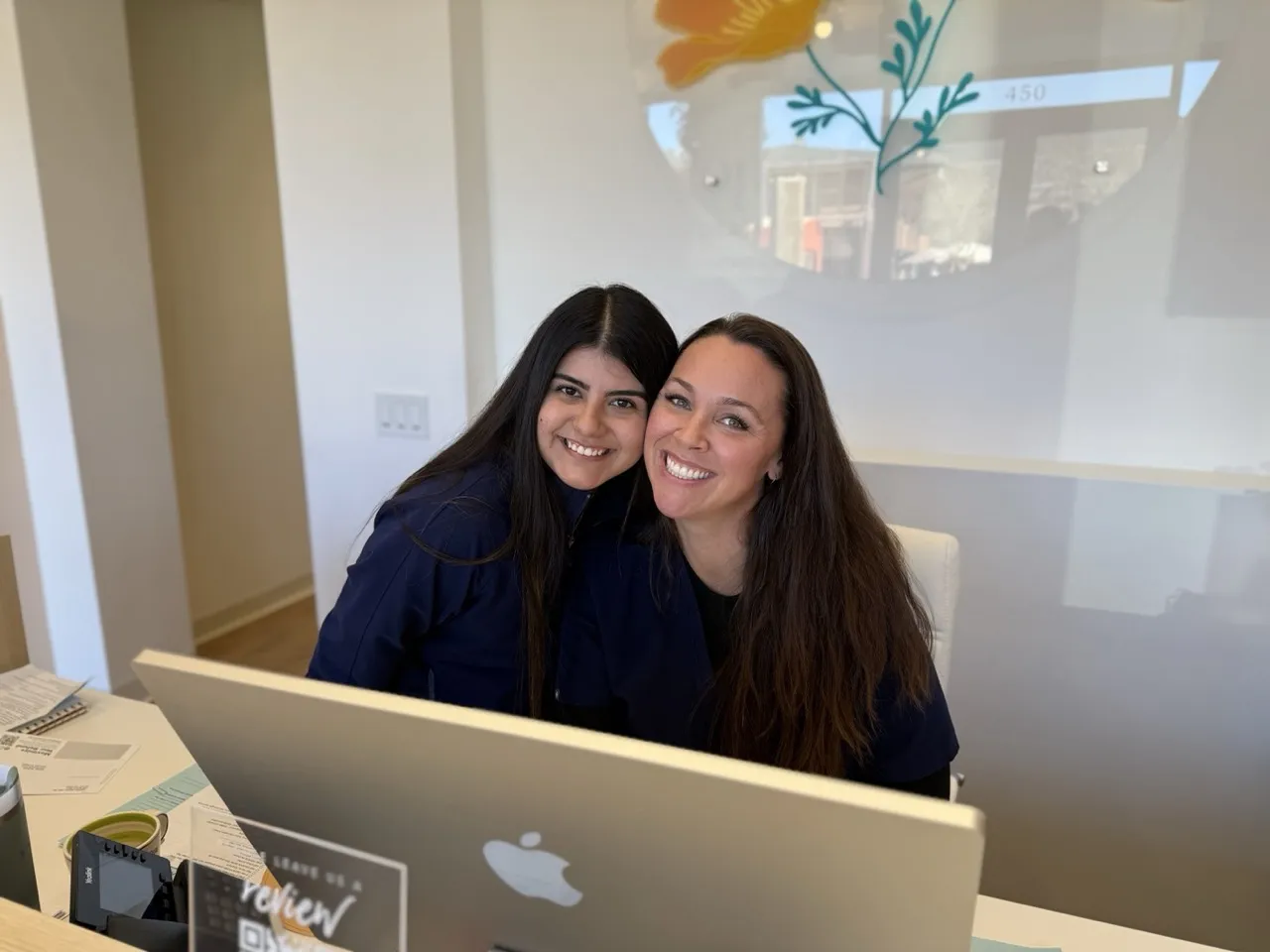 Two smiling women in dental scrubs sit behind a desk with a Mac computer in a bright, modern dental office. The wall behind them features a flower design and frosted glass