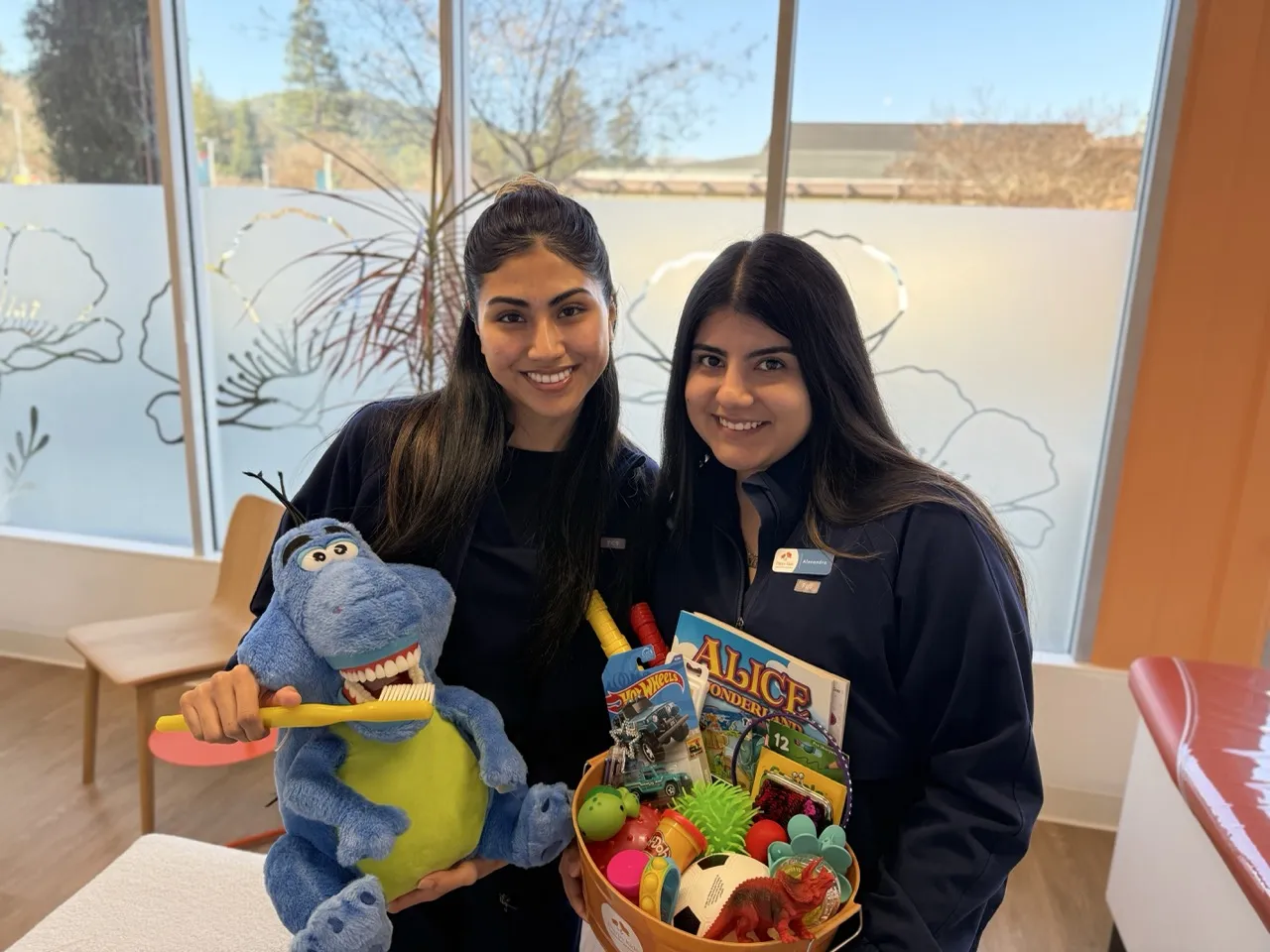 Two women in dental scrubs stand in a colorful pediatric dental office. One holds a plush dinosaur with a toothbrush, and the other holds a basket of toys and books, including "Alice in Wonderland.