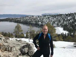 Dr.Andrea in athletic gear smiling, posing on a rocky outcrop with a snow-dusted pine forest and a lake in the background.