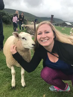 Dr. Andrea crouching and smiling next to a sheep in a green pasture with a rural farm landscape in the distance.